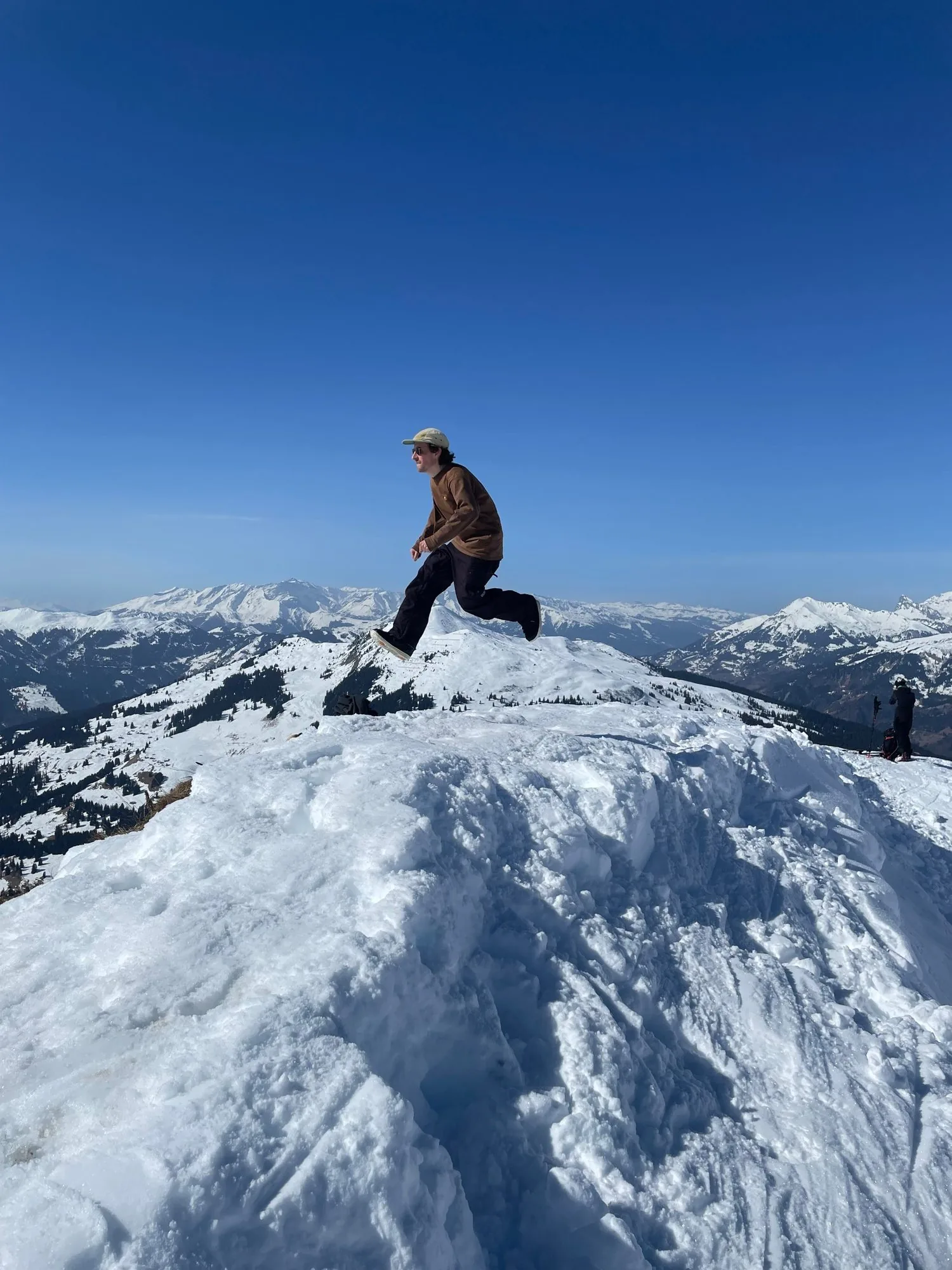 Frederic T. Kuntz mid-jump on a snowy mountain ridge under a deep blue sky