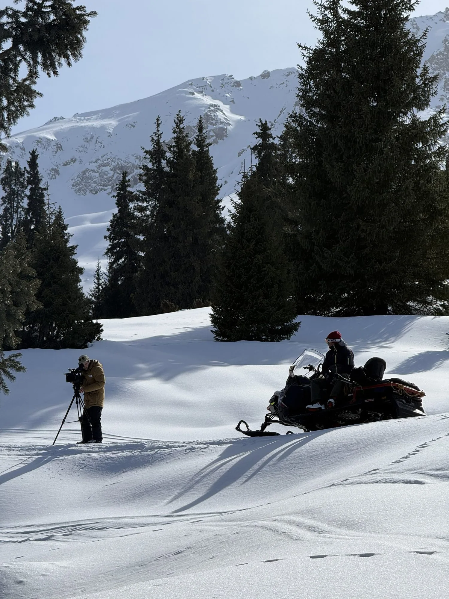Frederic T. Kuntz filming with a camera on a tripod in the snow, a snowmobile with a rider beside him